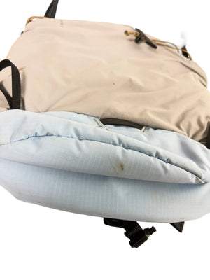 A close-up of a backpack showing its front panel with a light beige fabric section and a light blue padded lower section, with black straps and buckles visible.