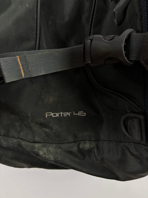 Close-up of a black Porter 46 backpack showing the brand name, a gray webbing strap with a black plastic buckle, and a small orange accent stitch on the strap.