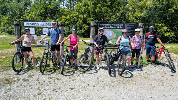 Group of cyclists posing with their bikes in front of a sign at a bike park entrance.
